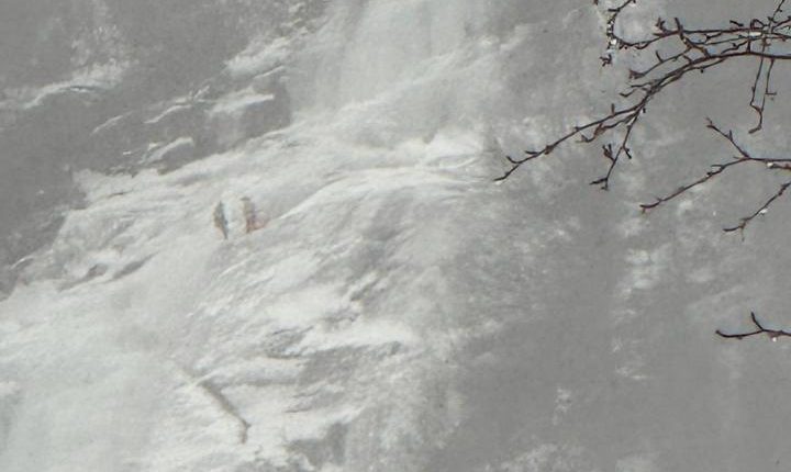 Snowy waterfall with people in the distance and bare branches in the foreground.