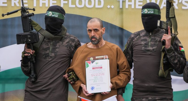 Israeli hostage Eli Sharabi, escorted by Hamas fighters, stands on a stage holding a document.