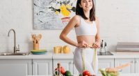 Selective focus of fit woman smiling at camera while measuring waist with tape near salad and fresh vegetables on table.