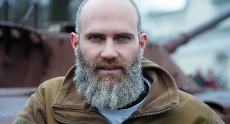 Portrait of a man with a long grey beard, wearing a tan jacket, near destroyed military vehicles.