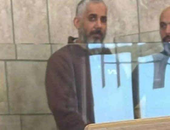 Man in brown shirt sitting behind a protective barrier in a courtroom.