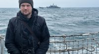 Journalist aboard a Royal Navy ship with a Russian vessel in the background.