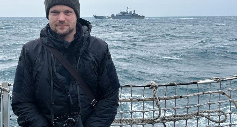 Journalist aboard a Royal Navy ship with a Russian vessel in the background.