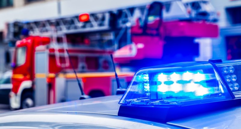 Close-up of a police car's flashing blue light with a firetruck in the blurry background.