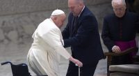 Pope Francis being assisted by two men, one in a suit and the other in clerical garb.