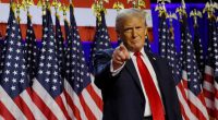Donald Trump pointing at the camera in front of American flags.