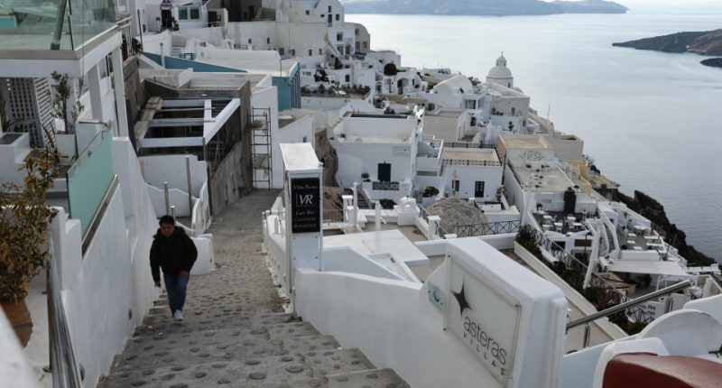 Person descending stairs in Santorini, Greece, with whitewashed buildings and ocean view.