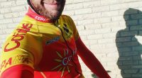 A cyclist in a yellow and red cycling jersey stands in front of a brick wall.