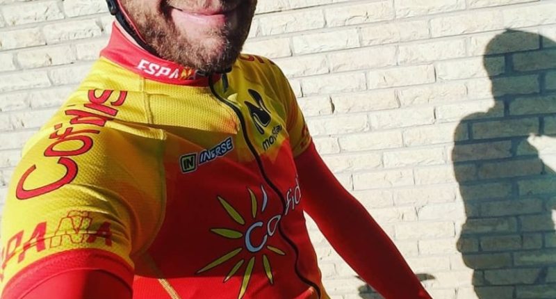 A cyclist in a yellow and red cycling jersey stands in front of a brick wall.