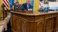 Elon Musk's son leaning on the Resolute Desk in the Oval Office as President Trump looks on.