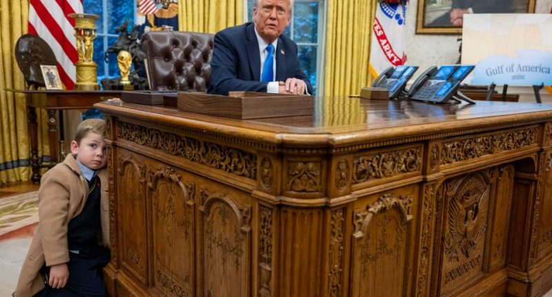 Elon Musk's son leaning on the Resolute Desk in the Oval Office as President Trump looks on.