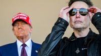 Elon Musk adjusting his sunglasses with Donald Trump in the background.