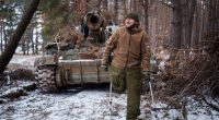 A smiling Ukrainian soldier, missing a leg, stands on crutches in front of a damaged self-propelled howitzer.