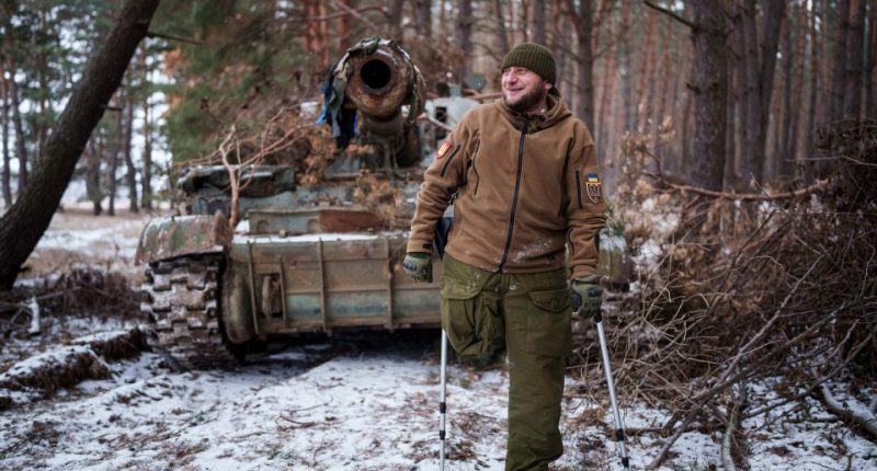 A smiling Ukrainian soldier, missing a leg, stands on crutches in front of a damaged self-propelled howitzer.