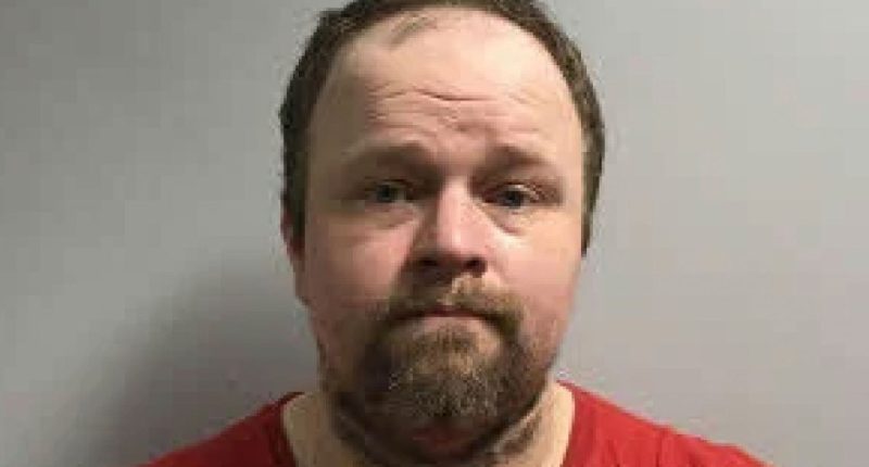 Mugshot of a man with a beard wearing a red shirt.
