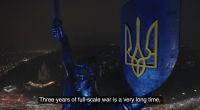Nighttime aerial view of a statue holding a shield with the Ukrainian trident, commemorating three years of war.