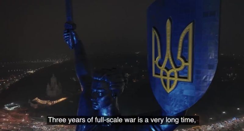 Nighttime aerial view of a statue holding a shield with the Ukrainian trident, commemorating three years of war.