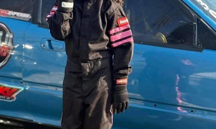 Girl in racing suit standing by blue car.