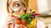 Woman using magnifying glass loupe, investigating shopping basket with many colorful vegetables. Healthy eating lifestyle, nutrients vegetarian food, searching for pesticides and chemicals.