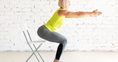 Yoga with a chair. Fit adult caucasian woman practice squat with props on a mat in loft white studio indoor, selective focus. Fitness, workout, trainer, sport, healthy lifestyle, concept.