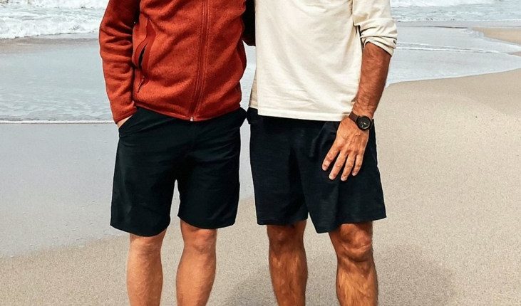 two men standing on a beach with one wearing sunglasses
