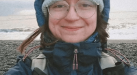 Selfie of a woman wearing glasses, a knit hat, and a dark blue jacket near the ocean.