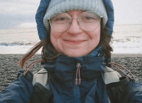 Selfie of a woman wearing glasses, a knit hat, and a dark blue jacket near the ocean.