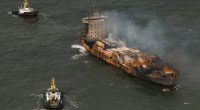 Aerial view of a burning cargo ship being towed by two tugboats.