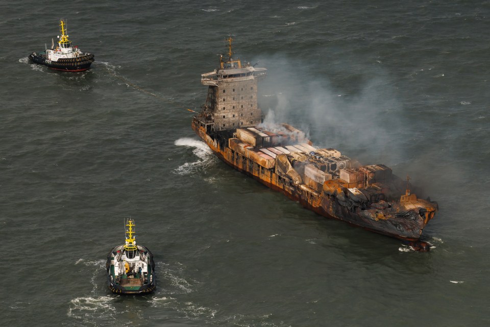 Aerial view of a burning cargo ship being towed by two tugboats.