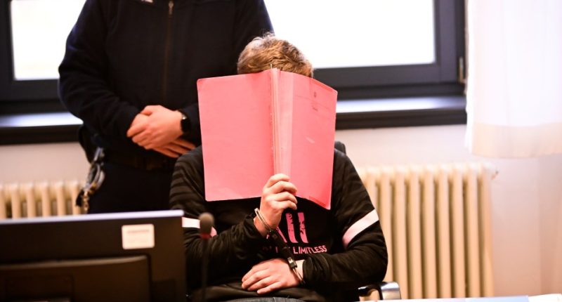 A man with his head down, partially obscured by a pink folder, stands in front of a uniformed officer.