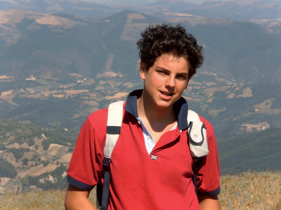 Carlo Acutis, a young boy, smiling while wearing a red shirt and backpack in a mountainous landscape.