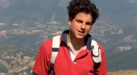 Carlo Acutis, a young boy, smiling while wearing a red shirt and backpack in a mountainous landscape.