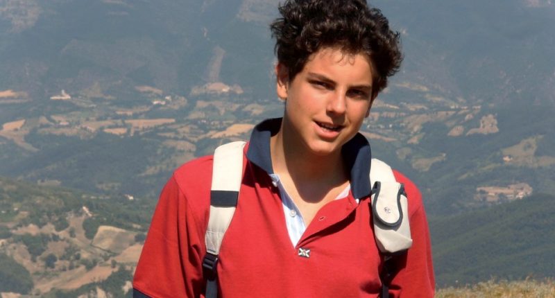Carlo Acutis, a young boy, smiling while wearing a red shirt and backpack in a mountainous landscape.