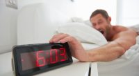 Man reaching for a digital alarm clock that reads 6:03.