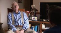 Elderly man in a blue shirt and suspenders being interviewed.