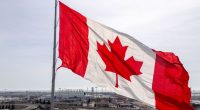 Canadian flag waving in the wind, with a factory and highway in the background.