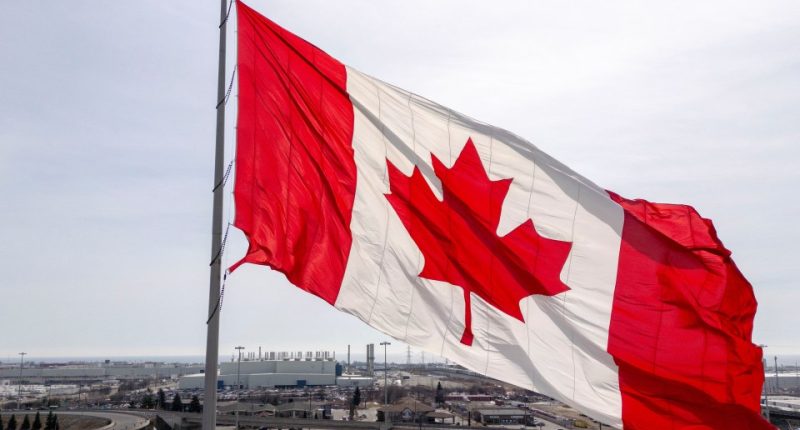 Canadian flag waving in the wind, with a factory and highway in the background.