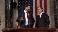 Two men in suits conversing at a podium in front of an American flag.