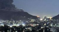 Nighttime view of a city with a large plume of smoke rising from a mountain in the background.