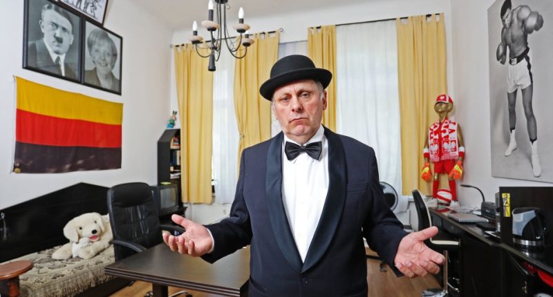 A man in a tuxedo and bowler hat stands with his arms outstretched in a room decorated with framed photos and a flag.
