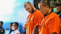 A British man in an orange prison jumpsuit stands with Indonesian police officers.