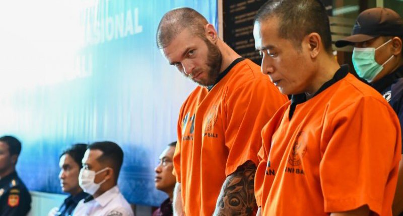 A British man in an orange prison jumpsuit stands with Indonesian police officers.