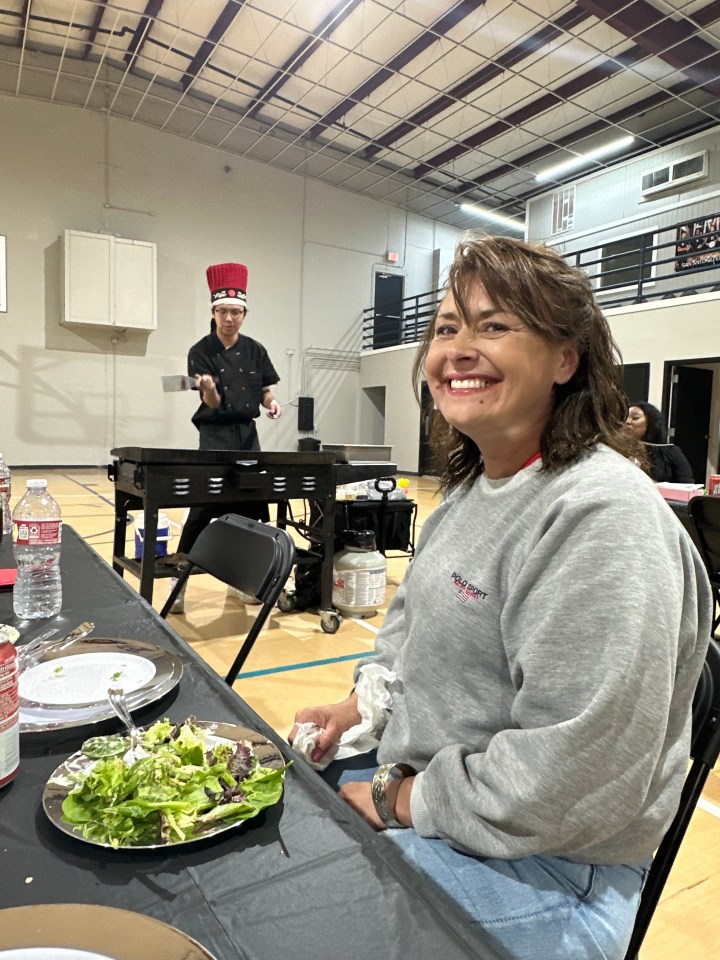 Woman smiles at the camera while sitting at a table with a salad in front of her; a chef prepares food in the background.