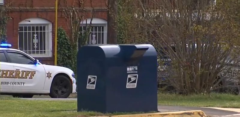 A blue USPS mailbox with a sheriff's car in the background.