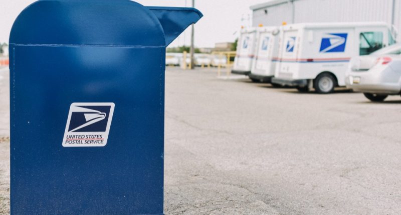 USPS postal box with USPS trucks in the background.