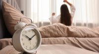 It's lazy morning o'clock. Alarm clock on bedside table and woman stretching in room, selective focus