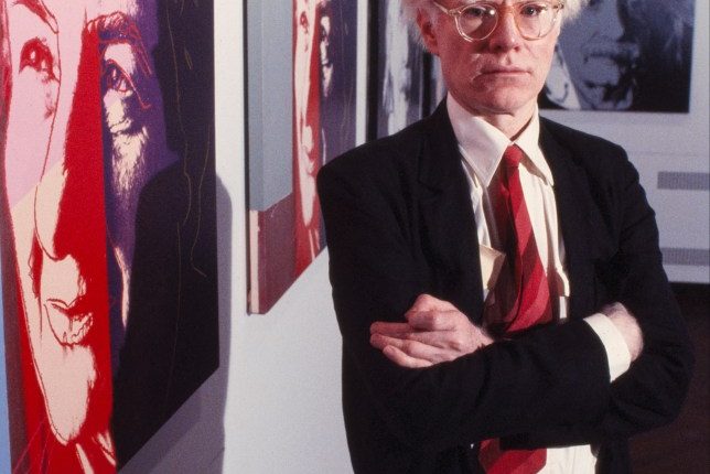 Andy Warhol standing in front of his artwork.