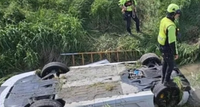 Upside-down supercar in a ditch with emergency personnel nearby.