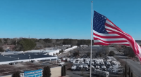Aerial view of a Camping World store with numerous RVs in the parking lot and a large American flag waving in the background.