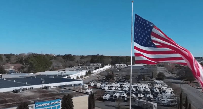 Aerial view of a Camping World store with numerous RVs in the parking lot and a large American flag waving in the background.